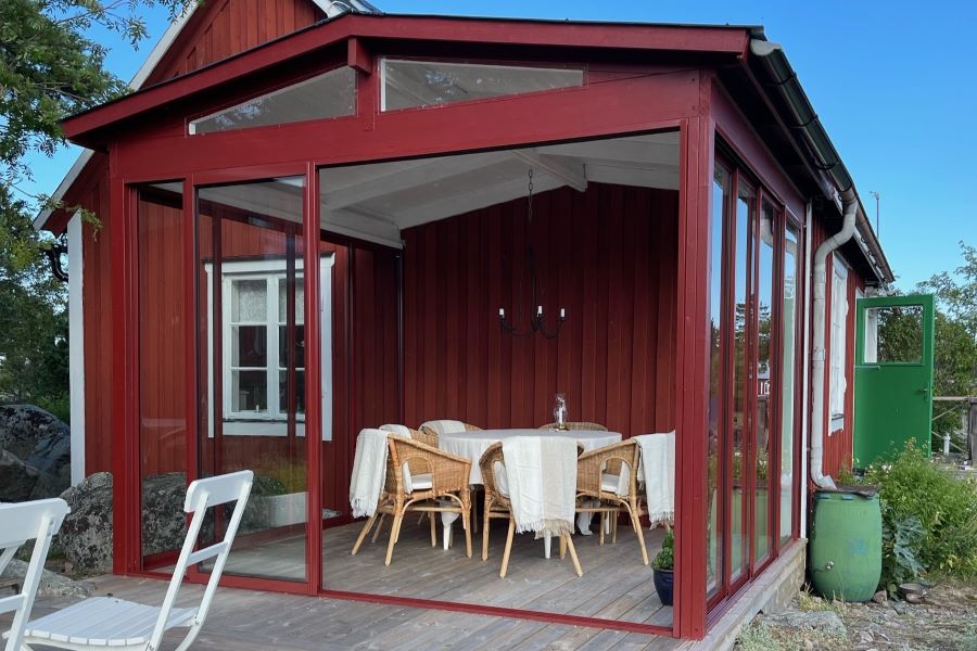 Ett charmigt falurött uterum med med skjutdörrar i glas som kan öppnas åt alla håll, vilket skapar en öppen och välkomnande känsla, beläget på en klippa med naturligt landskap och en grön dörr som öppnar mot en traditionell svensk trädgård. | Sommar M50 glaspartier | Mekoflex Uterum