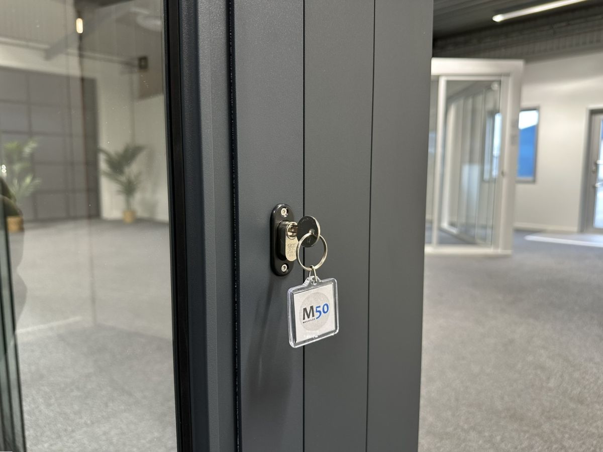 Hakregellås till skjutdörr i aluminium och glas, visat i närbild med nyckel. Bilden är tagen i en modern utställningshall för fönster och dörrar.
