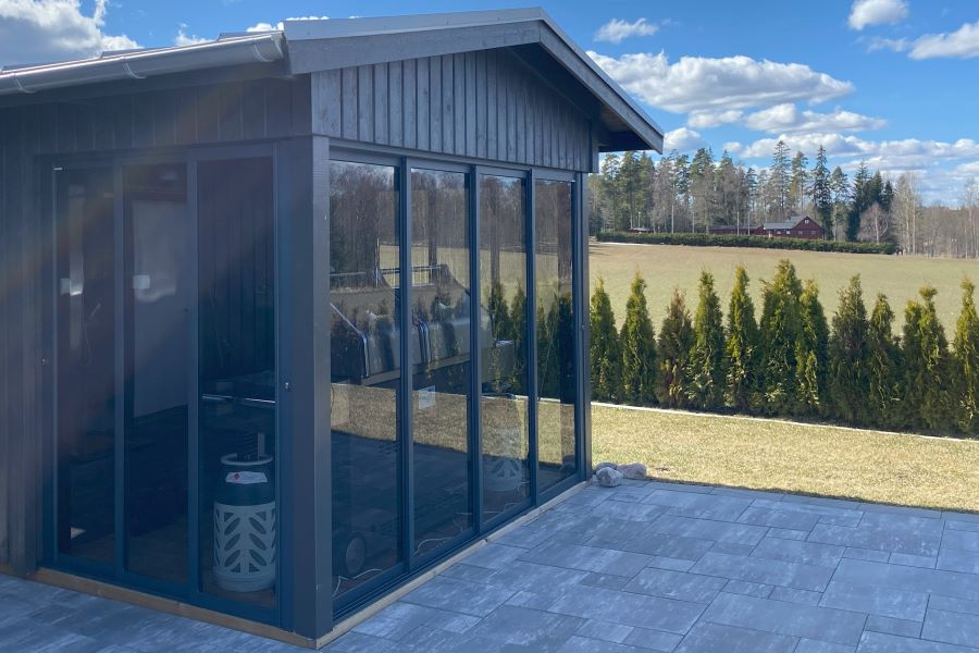 Modern uterumstillbyggnad med skjutdörrar av tonat glas som erbjuder utsikt över en lummig trädgård och ett fält | Sommar M50 glaspartier | Mekoflex Uterum