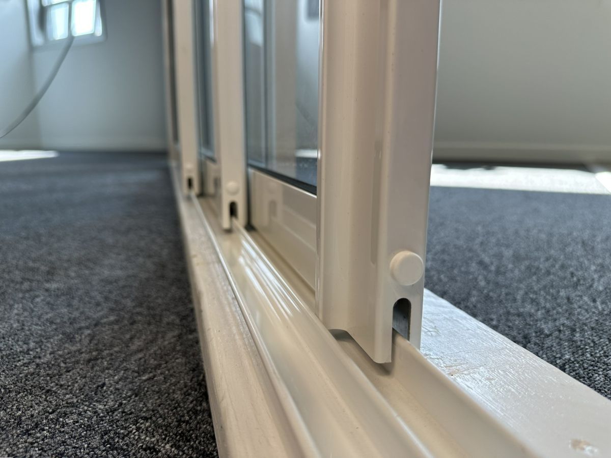 Närbild på undre rälsprofil med vita skjutdörrar till glasparti. Rälsen visar detaljerad konstruktion och hur skjutdörrarna löper smidigt längs spåret, anpassad för uterum.