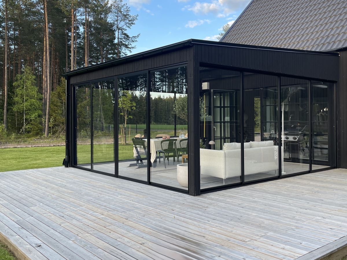 Elegant inglasat uterum med moderna svarta glaspartier och utsikt över en lummig skog. Inglasningen ger en ljus och rymlig känsla, vilket gör uterummet till en perfekt plats för avkoppling och middagar nära naturen.