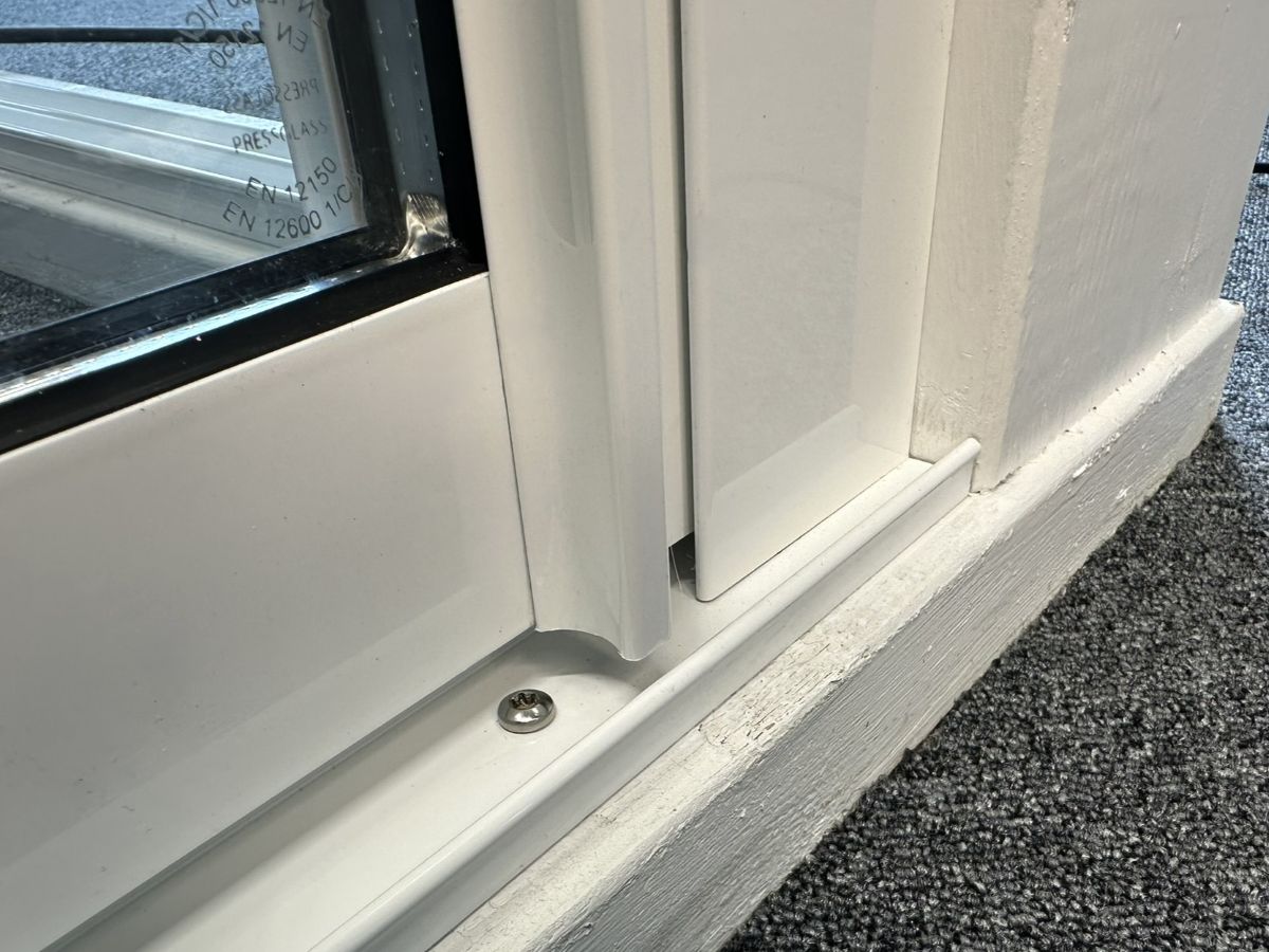 Närbild på undre räls och uppsamlingsprofil där en vit skjutdörr med integrerat handtag stängts. Måttanpassat glasparti med fokus på detalj och finish i beslag och karm.